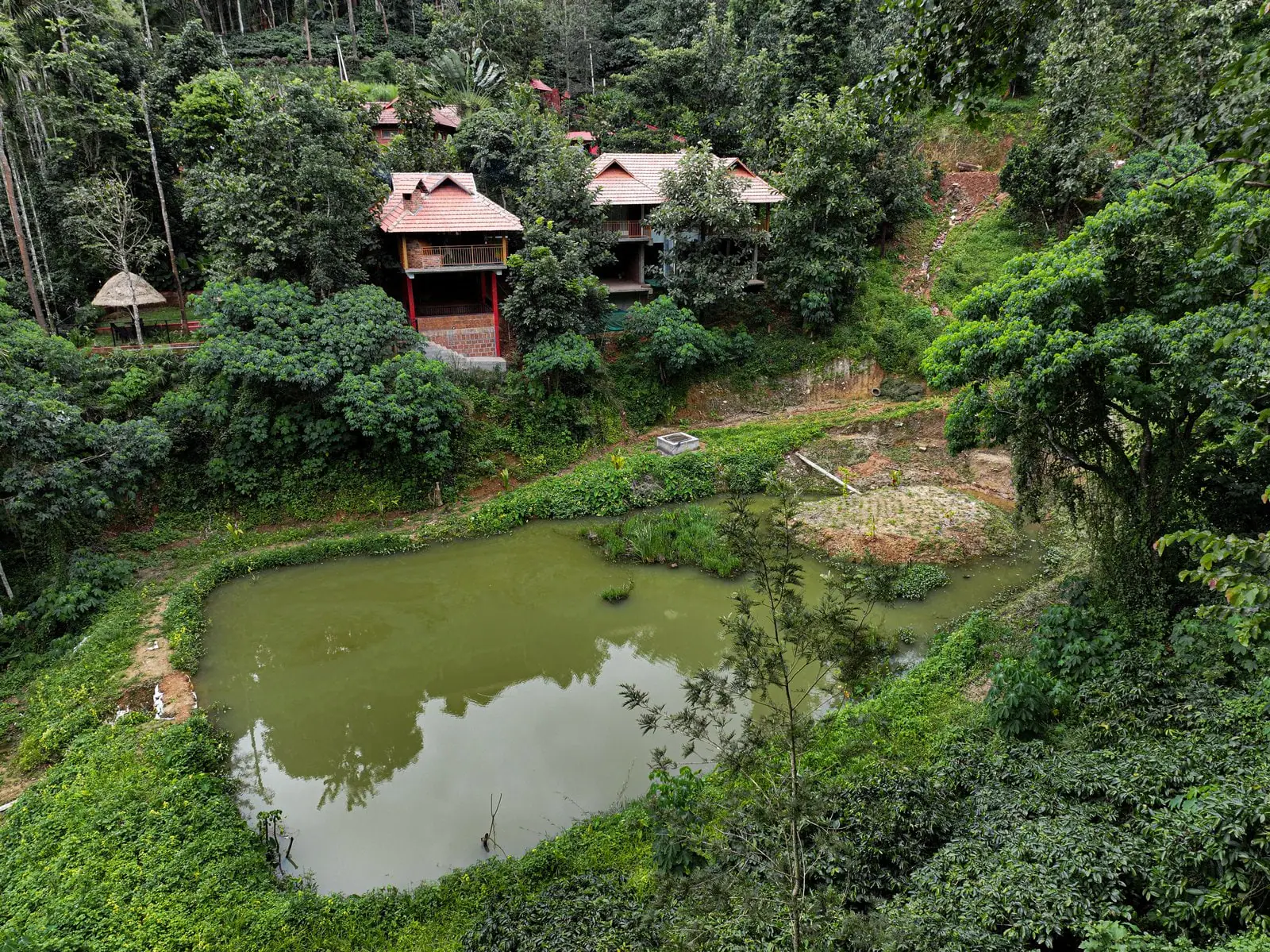 Two clay-tiled cottages with balconies raised on red posts, above a green pond in the estate's trees