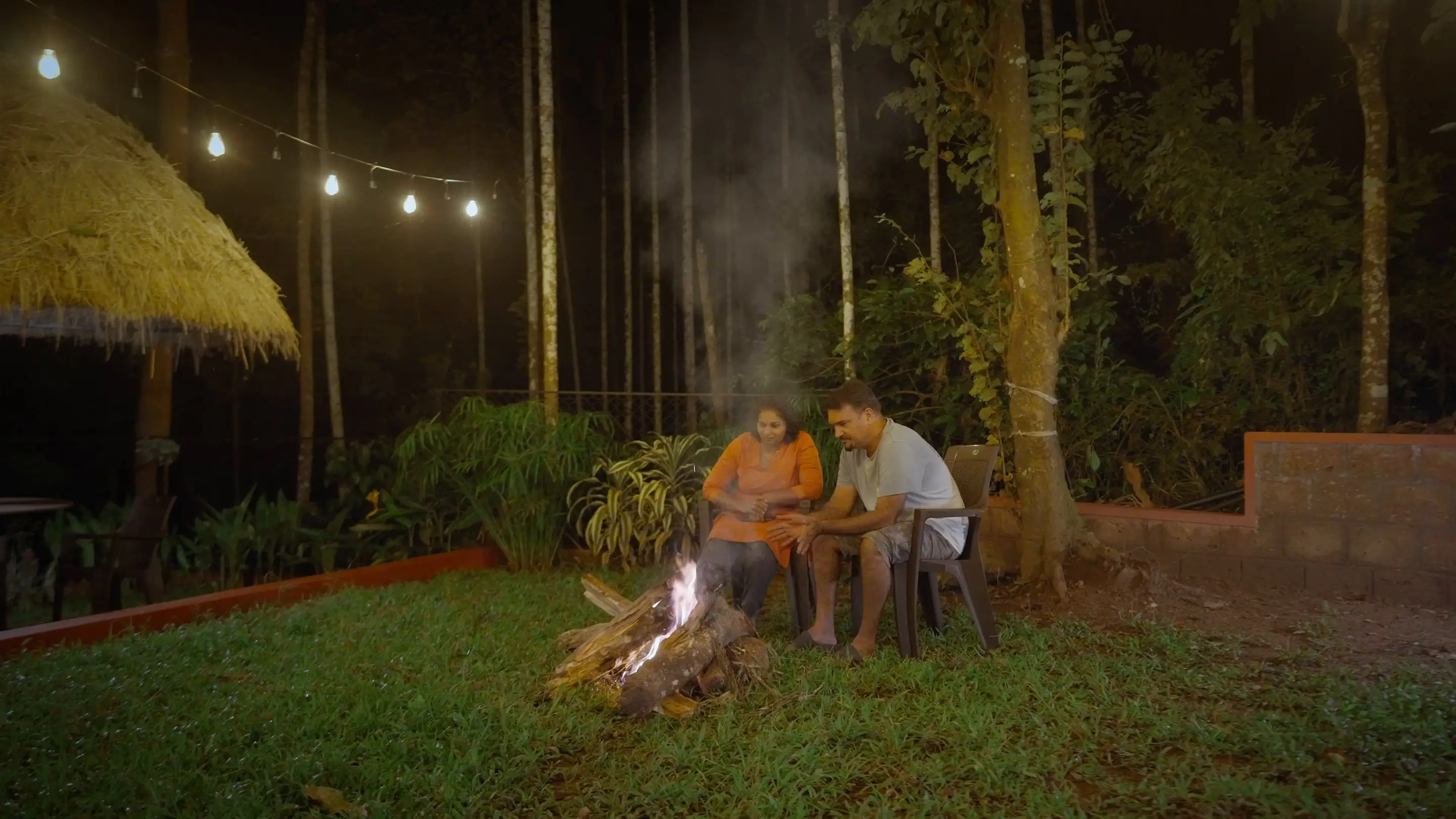 Two people sitting by a bonfire on the lawn at night, string lights strung through the trees above them