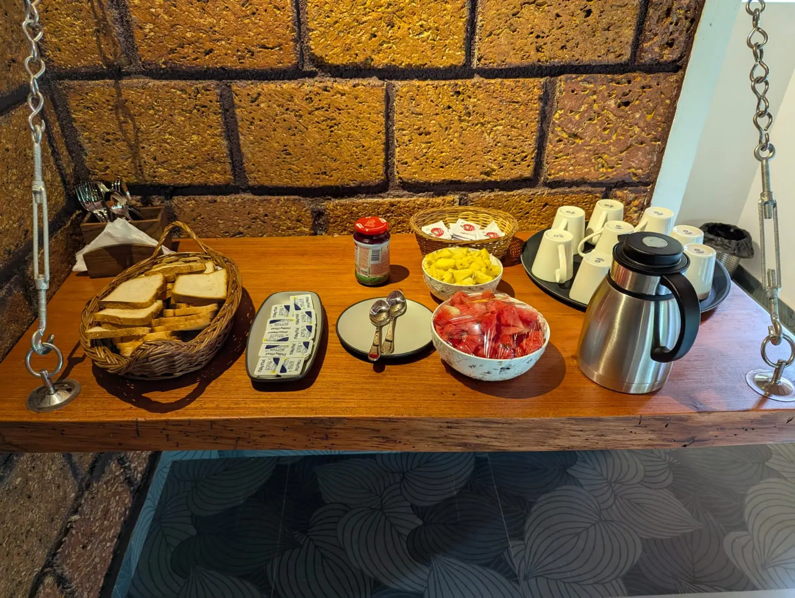 Breakfast on a timber counter against a laterite wall: a basket of bread, butter, bowls of cut fruit and a flask of coffee with cups