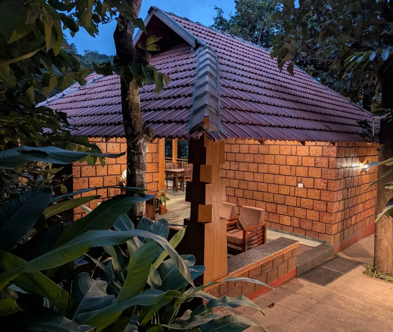 A laterite cottage under a clay-tiled roof at dusk, its lamps lit among the trees