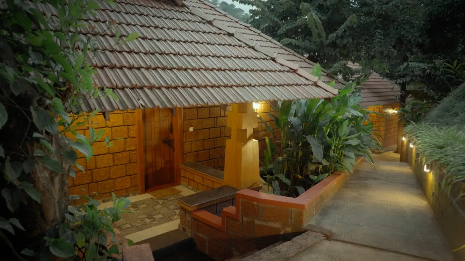 A laterite cottage with a clay-tiled roof and carved wooden door, a lamp-lit path running beside it at dusk