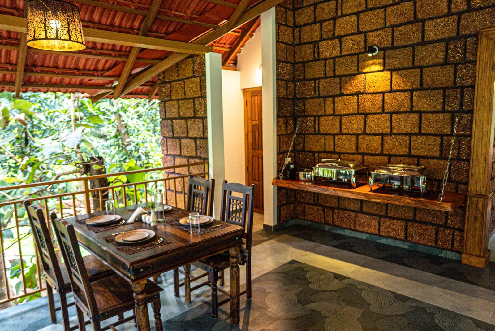 Chafing dishes on a timber counter hung on chains against a laterite wall, beside a laid dining table