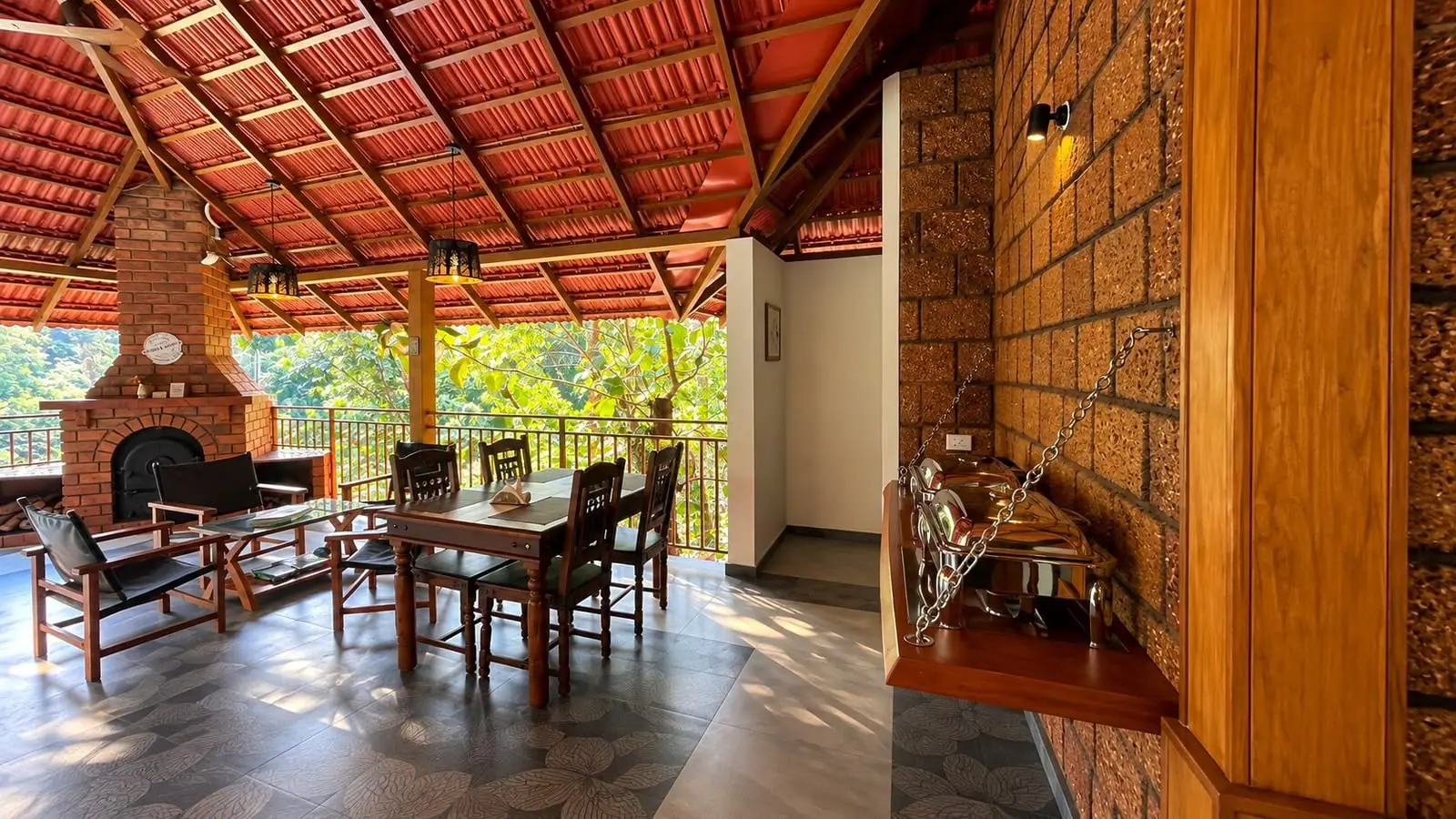 The dining pavilion by day: a tiled roof on timber beams, a laterite wall, a dining table and the brick fireplace, trees beyond the railing