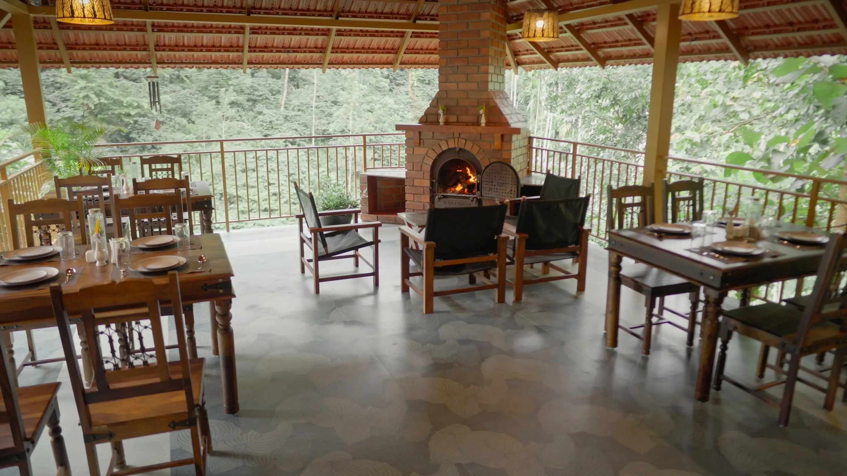 The open dining pavilion: timber tables laid for a meal around the brick fireplace, the forest beyond the railing