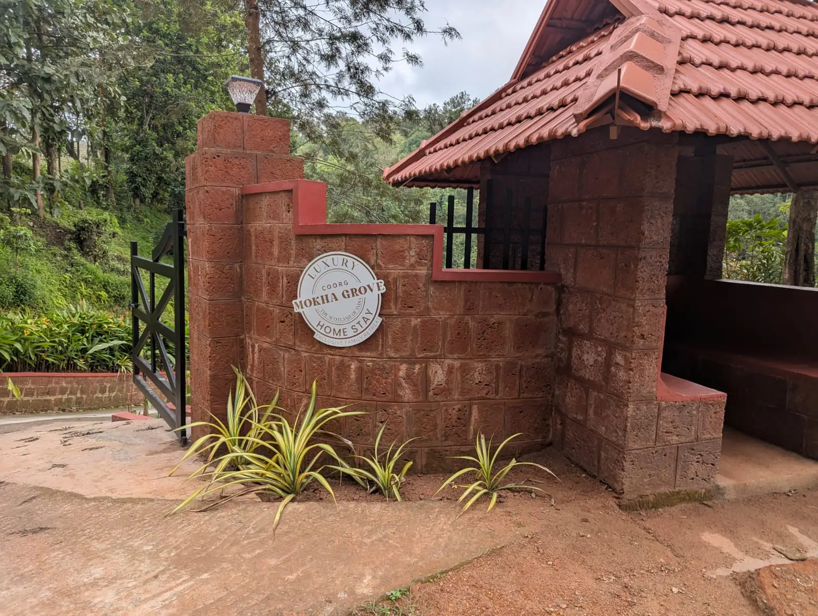 The estate gate: a curved laterite wall with a round Mokha Grove plaque beside a clay-tiled gatehouse