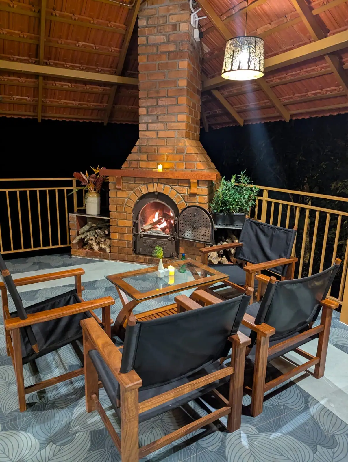 The pavilion's brick fireplace lit at night, logs stacked beside it and four timber chairs drawn up around a low glass table