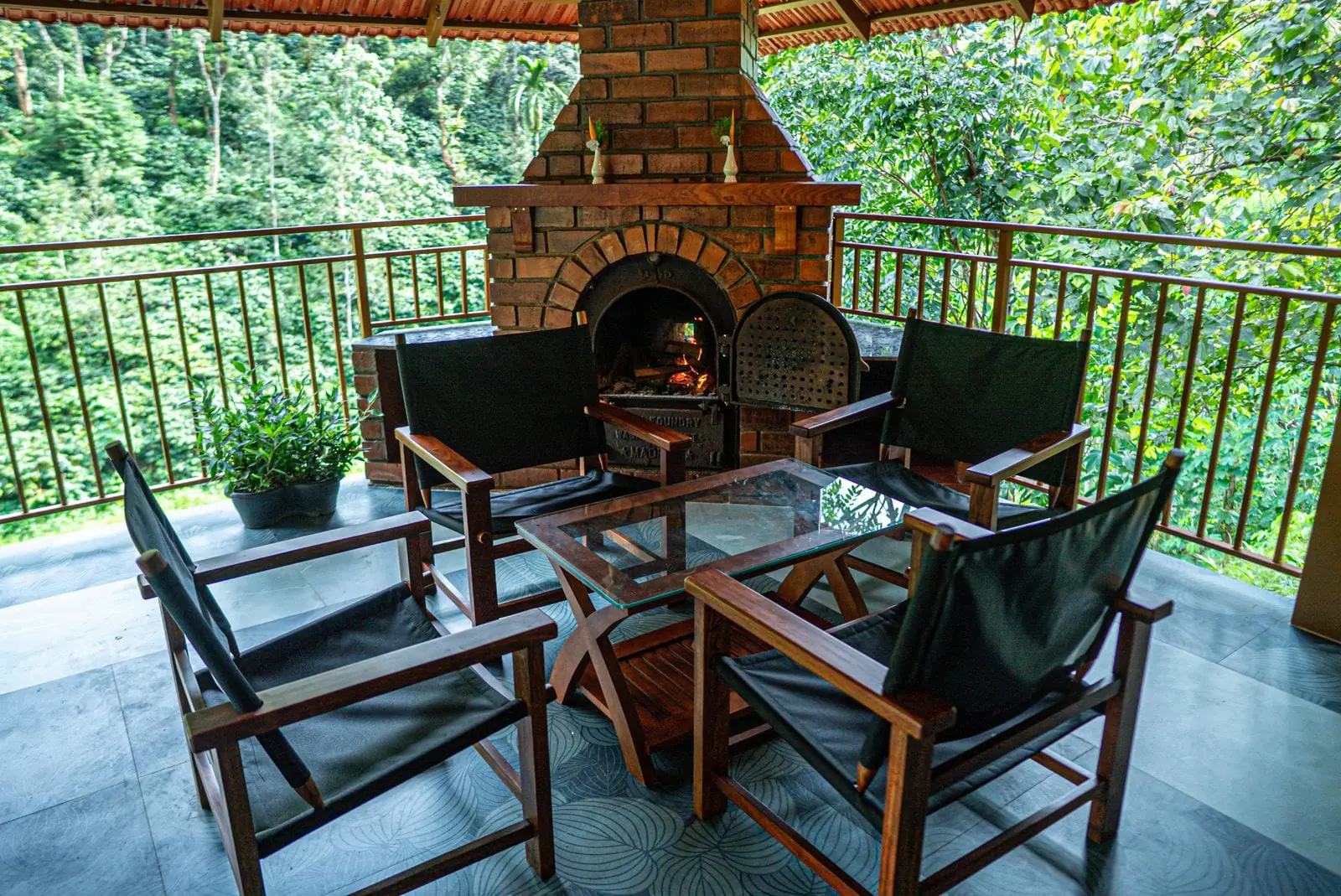 Timber chairs drawn up to a brick fireplace in the open pavilion, the forest beyond the rail