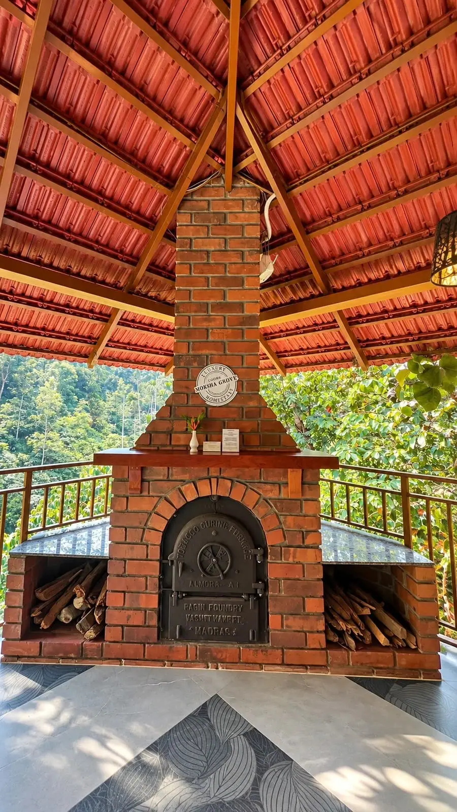 The brick fireplace in the open pavilion with the round Mokha Grove sign above it, trees beyond the rail