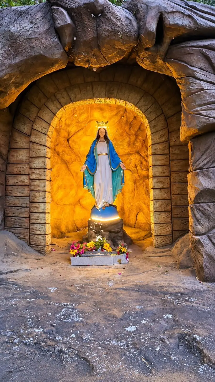 A statue of Mary lit warm gold inside a rock grotto, with fresh flowers at her feet