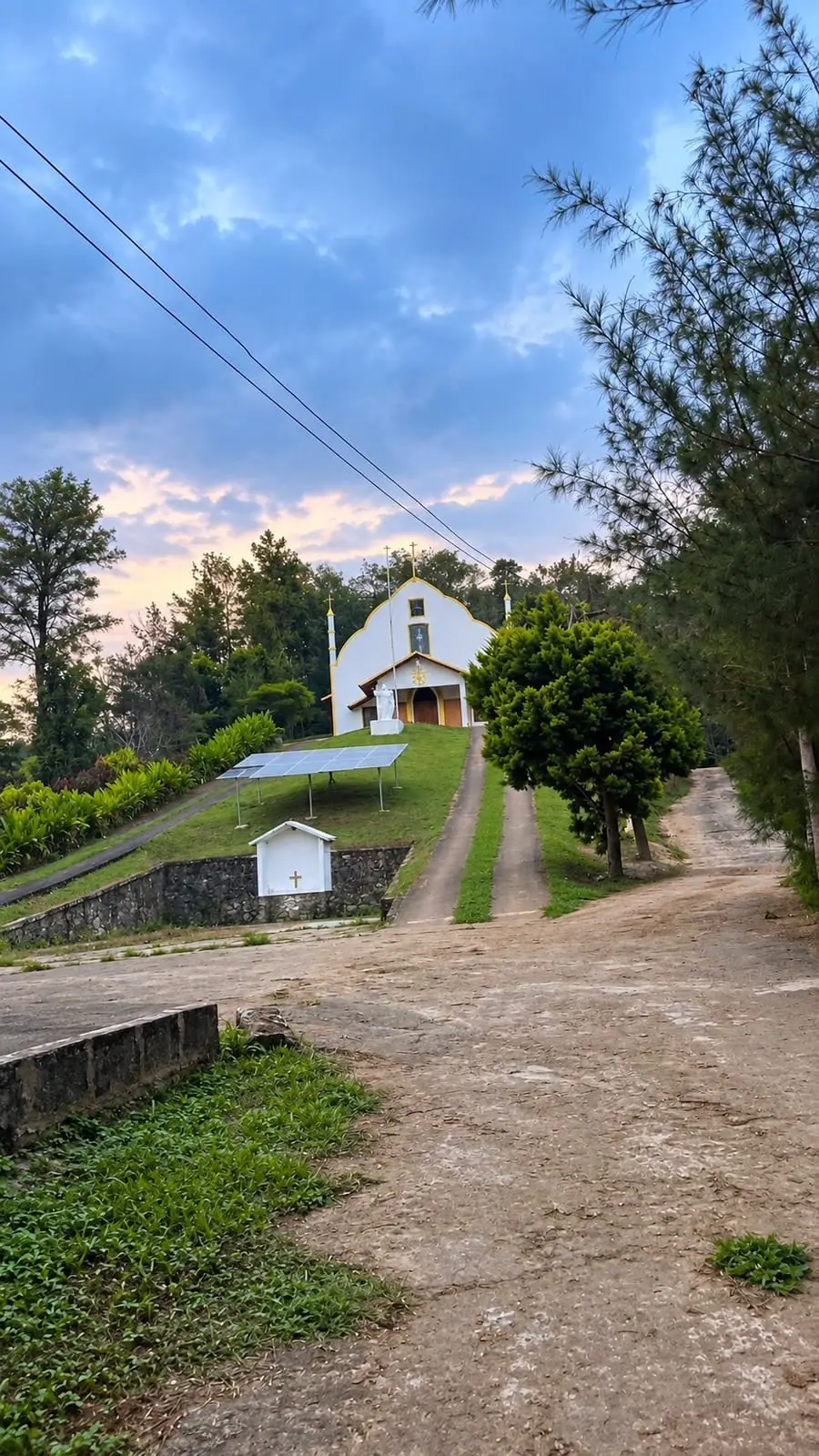 A white church with gold trim at the top of a grassy rise, a statue at its door and a track leading up through the trees under an evening sky
