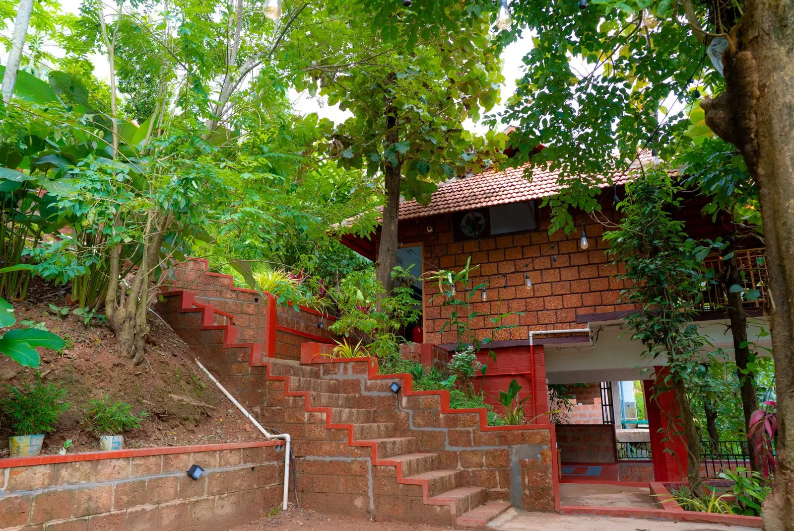 A clay-tiled building raised on red posts among tall trees, with a brick chimney and laterite steps below