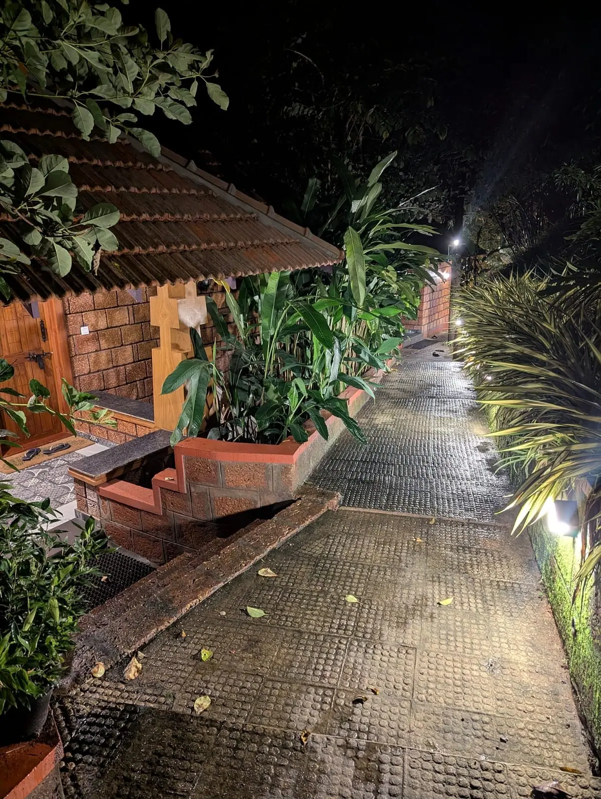 A cottage doorway under a clay-tiled roof at night, banana plants in a laterite planter and a lamp-lit path wet from rain
