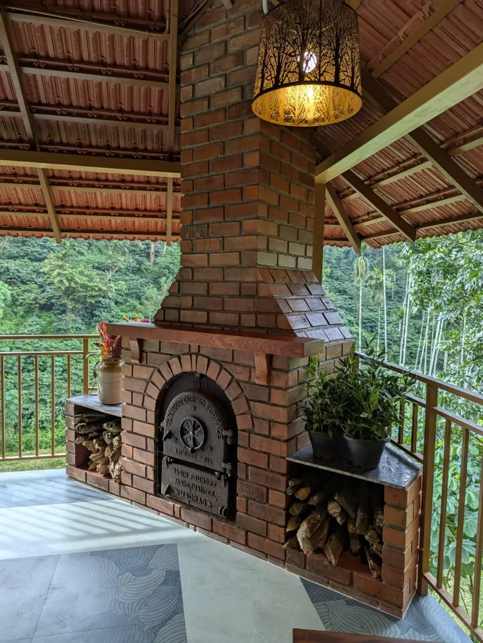 A brick fireplace with a cast-iron door in the open pavilion, plants beside it and trees beyond the rail