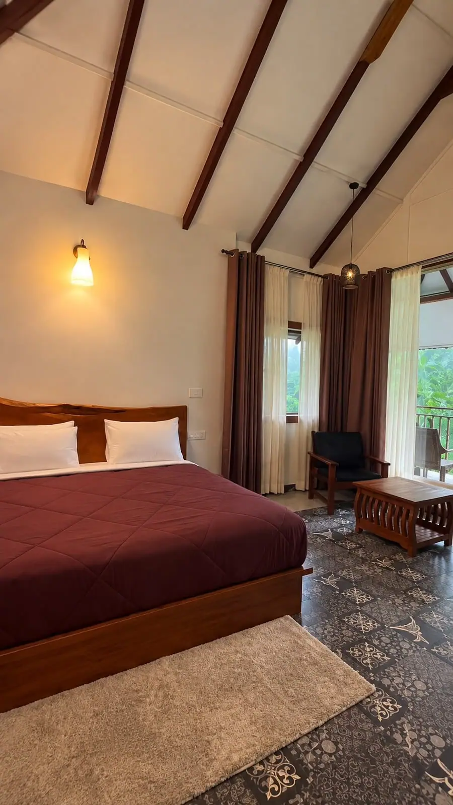 A bed with a deep red cover beside full-height curtains, trees outside the balcony door