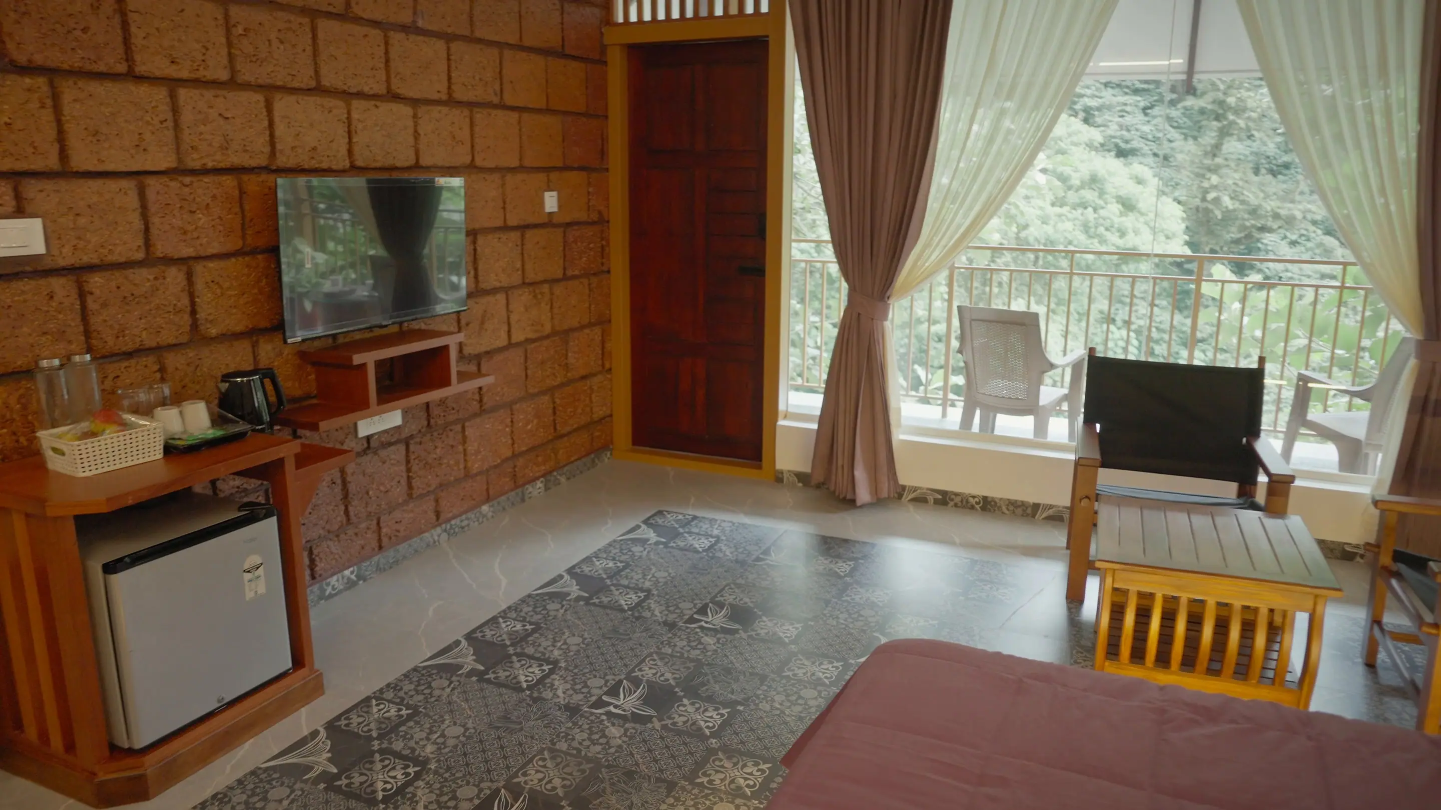 The suite's laterite wall with the kettle station, minibar and TV, the balcony door open to the trees