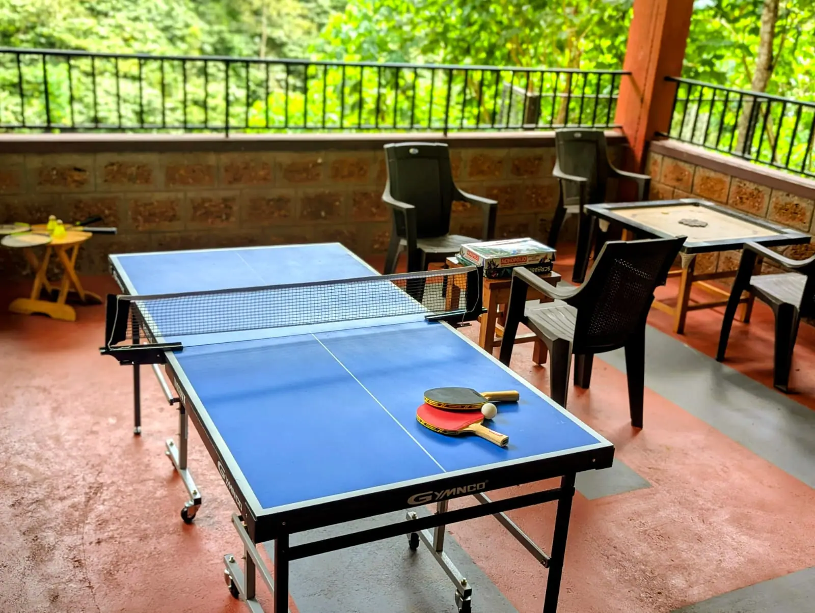 A blue table-tennis table with two bats and a ball on the red-floored veranda, chairs and a carrom table beyond